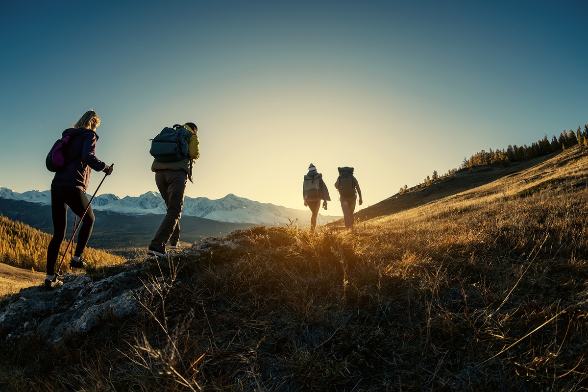 Four hikers with backpacks climbing a ridge towards the sunrise, snow-capped mountains behind